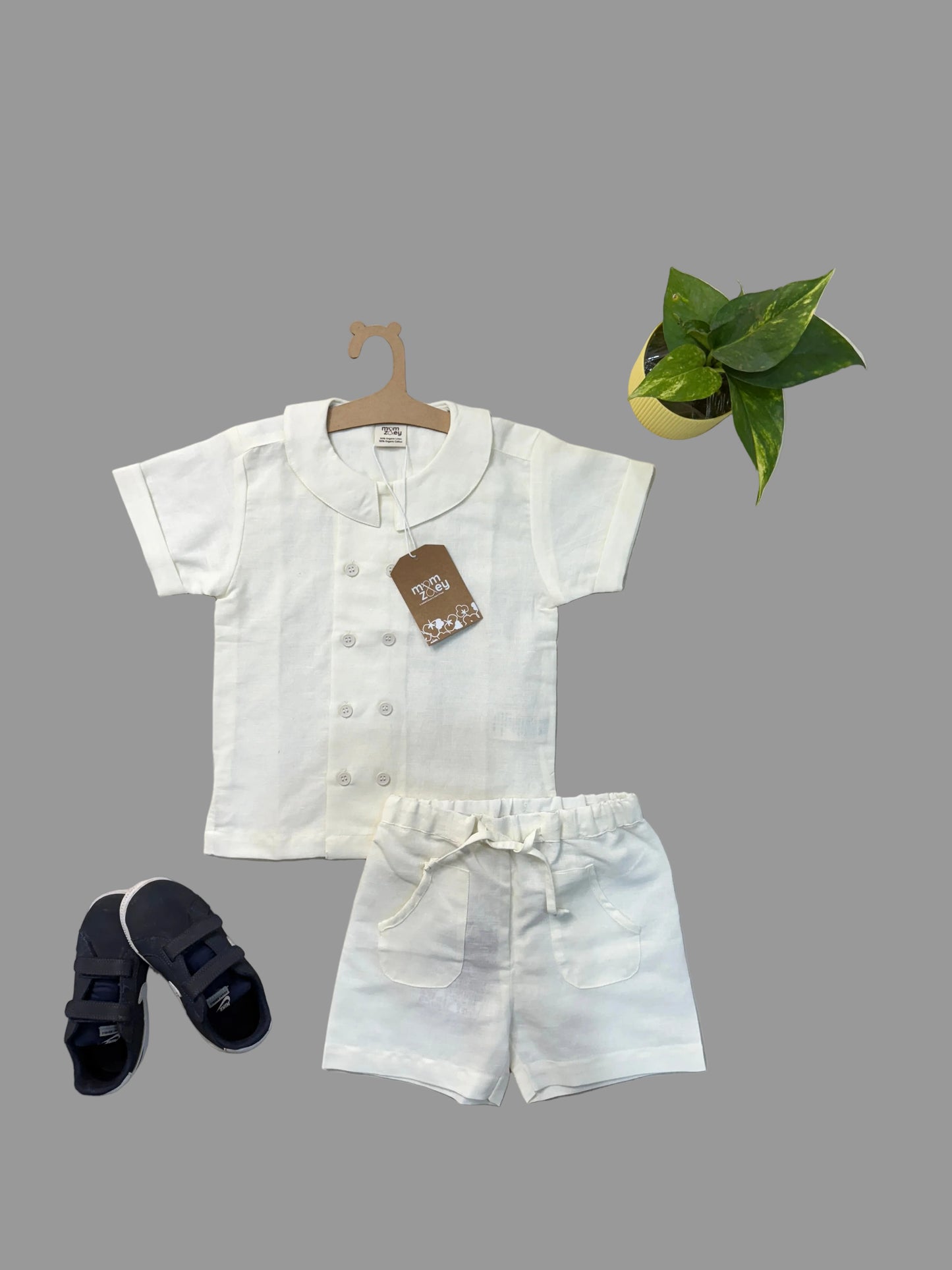 Children's white outfit with shorts on a hanger, accompanied by black sandals and a plant on a light gray background.